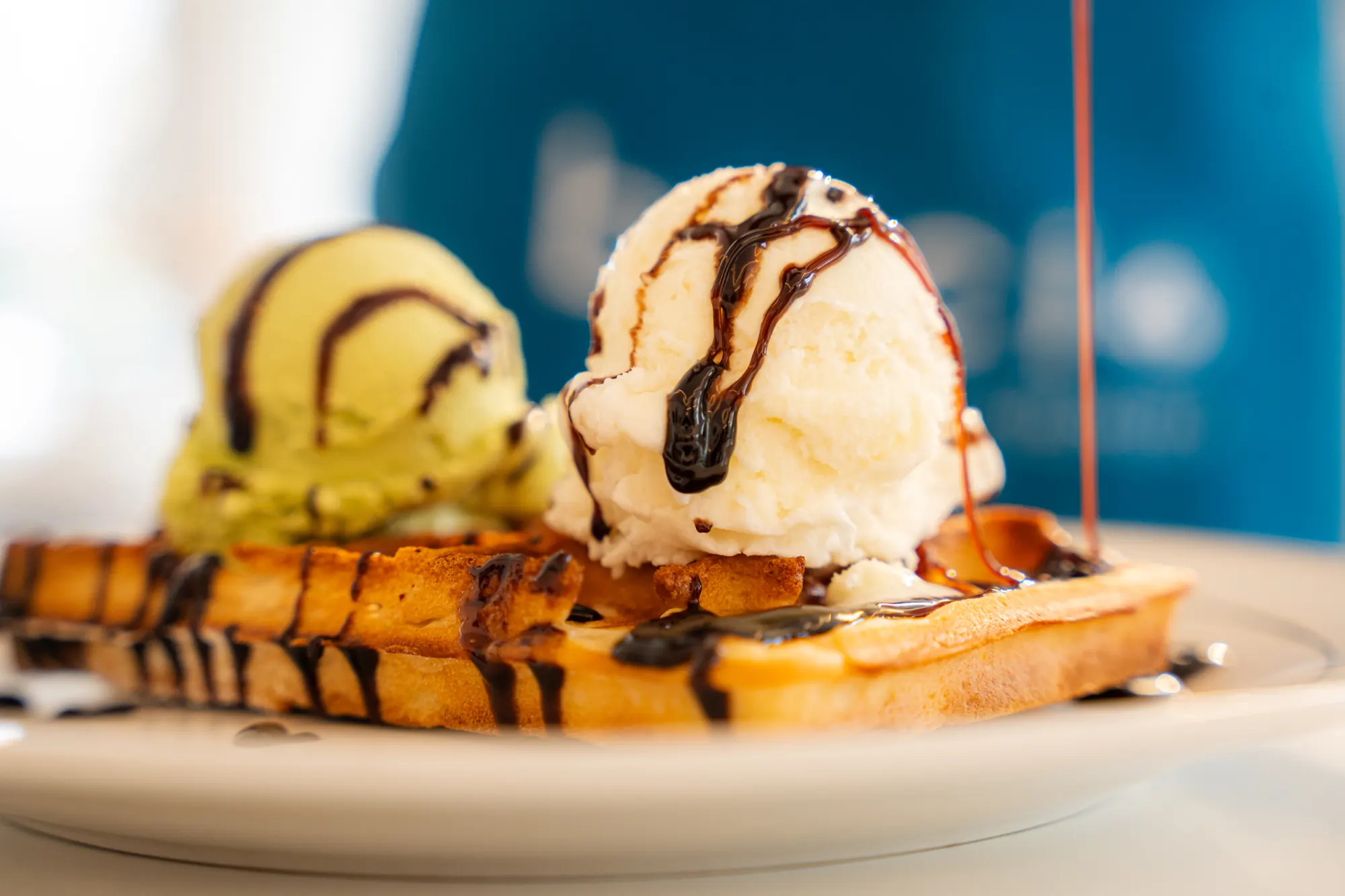 A waffle topped with pistachio and vanilla scoops under chocolate sauce