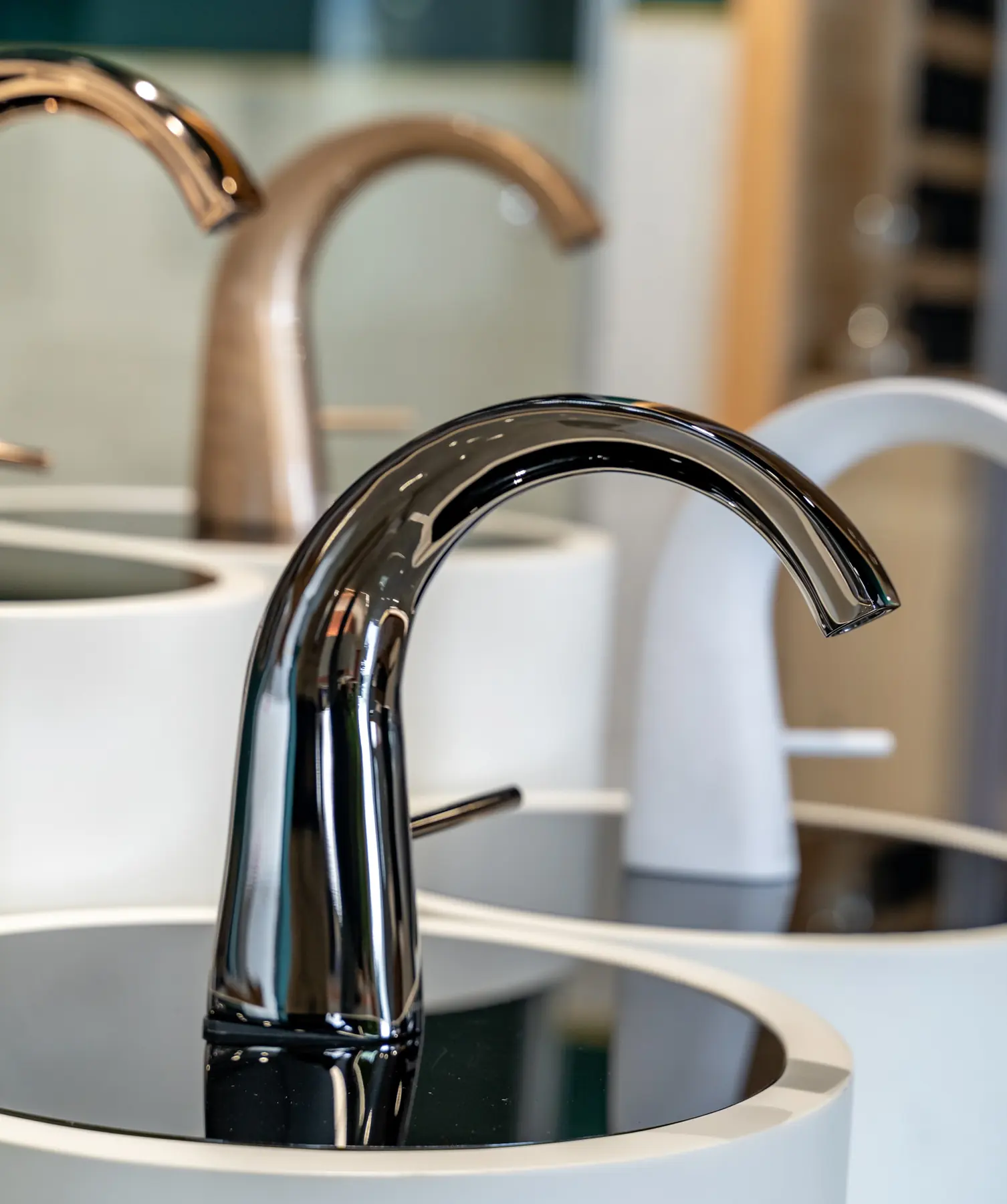A curved chrome faucet on a round basin, rose gold models behind