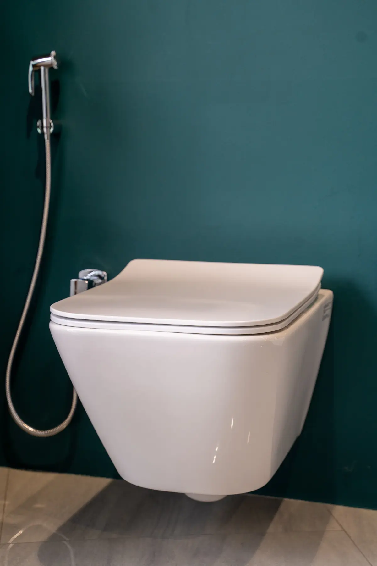 A wall-hung toilet and health faucet on a teal wall